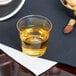 A clear hard plastic shot cup filled with liquid on a table with a bowl of nuts.