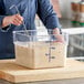 A woman using a metal utensil to mix a food mixture in a Choice clear square food storage container on a counter in a professional kitchen.