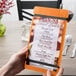 A person's hand holding a Menu Solutions wood menu board with rubber band straps on a table.