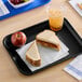 A black plastic fast food tray holding a peanut butter and jelly sandwich, an apple, and a cup of iced beverage.