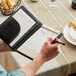 A man using the H. Risch, Inc. black vinyl server book to write at a table in a restaurant.