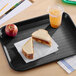 A black plastic fast food tray holding a peanut butter and jelly sandwich, an apple, and a cup of iced drink.
