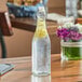 A clear glass bottle of water with a lemon slice and flowers on a table.