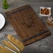 A customizable walnut wood menu board with rubber band straps on a table with a napkin, fork, and glass of whiskey and nuts.