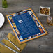 A Menu Solutions Country Oak wood menu clip board on a table with a fork and knife on a napkin.