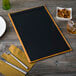 A customizable wood menu board with top and bottom strips on a table with a bowl of nuts and a glass of liquid.