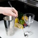 A hand holding a Barfly stainless steel Japanese style bar spoon next to a silver cup.