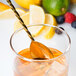 A Barfly gold plated bar spoon with a fork end stirring a glass of lemonade with ice and a lemon slice.