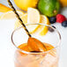 A Barfly gold plated bar spoon in a glass of liquid with a lemon slice.