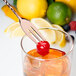 A copper plated Barfly bar spoon with a fork end stirring a glass with a cherry and lemons.