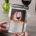 A hand holding a Menu Solutions Alumitique menu board with a glass of wine on a table.