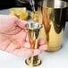 A person using a Barfly gold-plated Japanese style jigger to pour liquid into a gold cup.