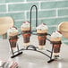 An Acopa metal flight carrier holding four flared Pilsner glasses filled with brown and white layered desserts.