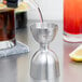 A person using a Barfly stainless steel bell jigger to pour a red and white liquid into a silver metal cup.