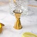 A gold-plated Barfly Japanese style jigger next to a glass and a slice of lemon.