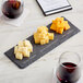 Acopa black slate tray with cheese cubes and wine on a table.