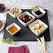 A black slate tray with small white bowls of food and a knife on a table.