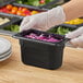 A person in gloves holding a black Cambro food container filled with shredded cabbage.