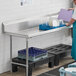 A man in a blue shirt standing at a stainless steel Regency clean dish table with dishes on a rack.