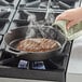 A hand using a green pot holder to hold a Valor cast iron grill pan with a steak cooking on it.