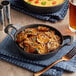 A Valor pre-seasoned cast iron round casserole dish filled with cheesy potatoes next to a glass of beer on a table.