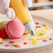 A hand using an Ateco plain piping tip to pipe cream onto a pink macaroon.