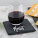 A glass of wine on a slate plate next to cheese with a slate coaster that says "merlot"