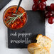 A black slate plate with red pepper bruschetta, bread, and a bowl of food with a rosemary sprig on a counter.