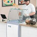 An Avantco flat glass sneeze guard on a counter with a glass case.