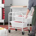 A man in a chef's uniform pushing a Regency mobile aluminum lug rack with food in it.