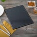 A black Menu Solutions Hamilton menu on a wood table with silverware.