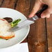 A person uses a Walco Denver Chop steak knife with asparagus on a plate to cut a piece of meat.