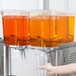 A hand pouring orange juice into a clear plastic container from a Crathco double beverage dispenser bowl.