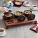 A person putting food into a Valor mini cast iron pot on a table with a pan of food.