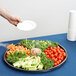 A person holding a black round catering tray filled with vegetables.