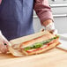 A person in gloves holding a sandwich in a Bagcraft Dubl View ToGo! Bakery Bag on a cutting board.