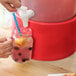 A hand filling a glass with a straw using a white plastic faucet on a Choice red beverage dispenser base.