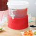 A red Choice beverage dispenser base on a table with drinks and fruit.