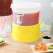 A yellow Choice beverage dispenser base on a wooden surface with drinks and fruit.