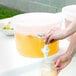 A person pouring a liquid into a clear round beverage dispenser.