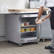 A man in a white shirt and black pants putting a container of chicken into a Beverage-Air undercounter refrigerator in a school kitchen.