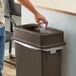 A person putting a plastic bottle into a brown Lavex slim rectangular trash can with a Drop Shot lid.