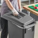 A woman in plastic gloves and an apron putting a lid on a Lavex grey rectangular trash can.