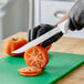 A 9-inch serrated edge utility knife with a white handle being used to slice a tomato on a green cutting board.