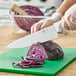 A 10-inch chef knife with a white handle being used to slice a head of purple cabbage on a green cutting board.