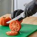 A 6-inch serrated edge utility knife with a white handle being used to slice a tomato on a green cutting board.