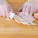 A person in gloves uses a Choice 6" Wide Stiff Boning Knife to cut up chicken on a wooden table.