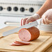 A 12-inch Granton edge slicing knife with a white handle being used to slice ham on a wooden cutting board.