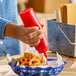 A red plastic squeeze bottle being used to dispense sauce onto a tray of curly fries.