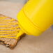 A person using a yellow Choice wide mouth squeeze bottle to pour mustard on a piece of bread.